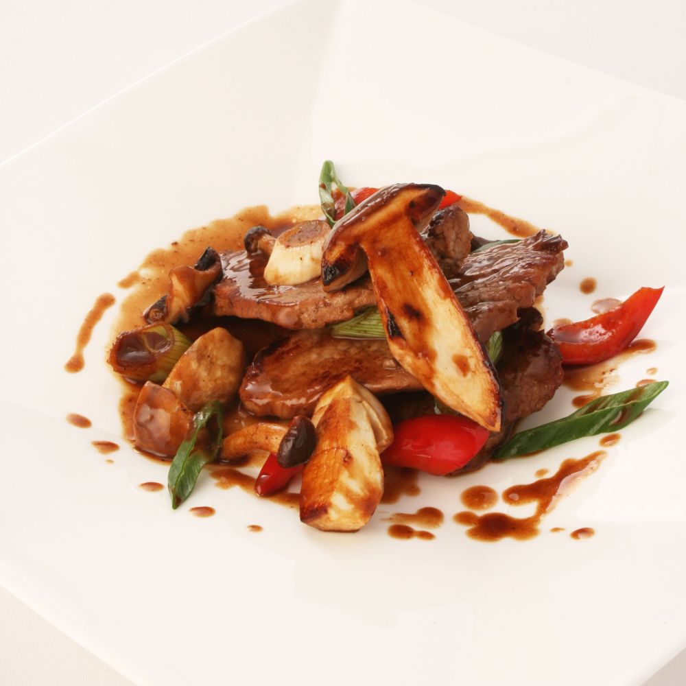 Ein kunstvoll angerichtetes Gourmetgericht mit gegrilltem Fleisch, Garnele, Zucchinistreifen und frischen Kräutern auf einem weißen Teller, serviert von einem Kellner in einem Restaurant.