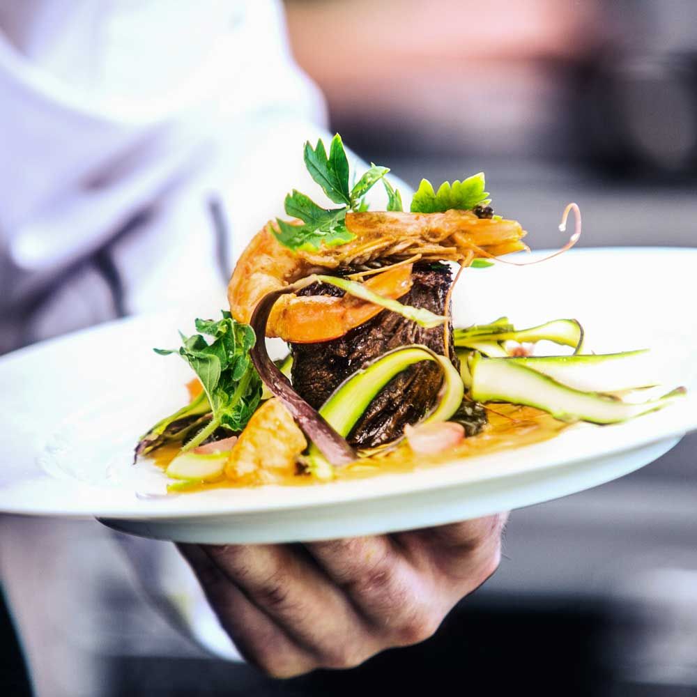 Ein kunstvoll angerichtetes Gourmetgericht mit gegrilltem Fleisch, Garnele, Zucchinistreifen und frischen Kräutern auf einem weißen Teller, serviert von einem Kellner in einem Restaurant.